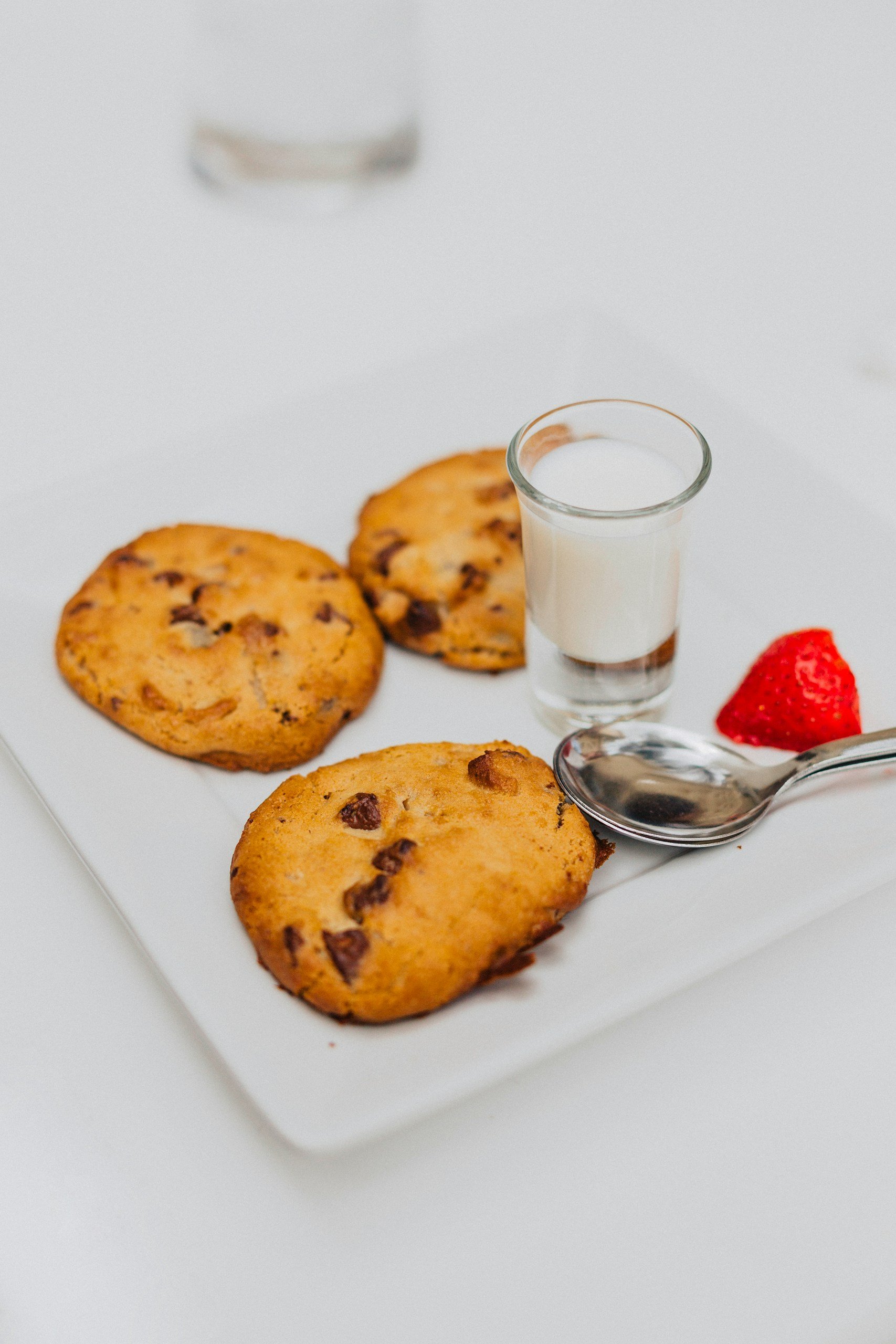 Plate with three chocolate chip cookies, a cup of milk, and a fresh strawberry.